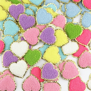 Op de afbeelding: Een verzameling hartvormige patches in verschillende kleuren, waaronder roze, paars, blauw, groen, geel en wit. Elk hart heeft een gouden rand. De patches hebben een fuzzy textuur.
