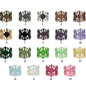 May include: A collection of nineteen ornate metal ring blanks in various colors. Each ring features an openwork filigree design and a circular setting for embellishments. Colors include bronze, black, silver, gold, and shades of green, blue, and pink.