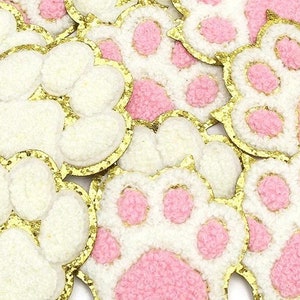 Op de afbeelding: Close-up van verschillende witte en roze pootvormige patches met gouden glitterranden. De voetzolen zijn roze, de rest is wit. Deze patches zijn ideaal voor knutselen en geven diverse projecten een schattige uitstraling.