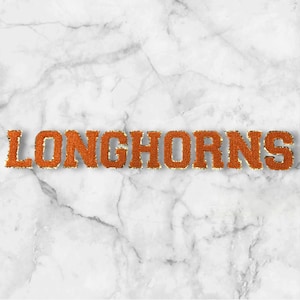 Könnte beinhalten: Das Wort "LONGHORNS" in orangefarbenen, strukturierten Buchstaben mit goldenem Rand, vor einem weißen und grauen Marmorhintergrund. Die Buchstaben sind fett und stechen hervor.