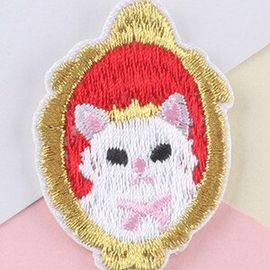 May include: Embroidered patch of a white cat with black eyes and a pink bow, set against a red background. The cat is framed by a gold, ornate border, creating a decorative and whimsical design.