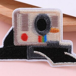 May include: Embroidered patch of a vintage Polaroid camera. The patch has a black and white body with a red dot, a rainbow stripe, and a black lens. The patch is edged with white stitching.