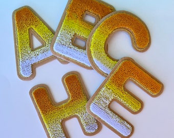 May include: A collection of embroidered fabric letters in the shape of A, B, C, F, and H. The letters feature a gradient of colors, transitioning from gold to yellow to white. Each letter is outlined in gold and has a tan backing.