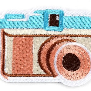 May include: Embroidered patch of a retro camera. The patch has a light blue top, a peach and cream body, and a brown lens. The patch has a white border and is designed for application on clothing or accessories.