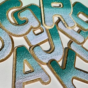 May include: Close-up of embroidered fabric letters in shades of teal and white, with gold trim. The letters are scattered on a white surface. The letters appear to be patches or appliques, suitable for crafts or personalization.