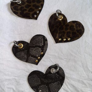 Peut inclure: Quatre breloques en forme de cœur en matériau texturé. Deux présentent un imprimé léopard marron et noir avec des œillets métalliques argentés ou dorés et des petits clous. Deux présentent un motif peau de serpent noir avec des œillets.