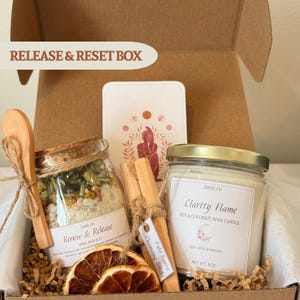 Puede incluir: Una caja Release & Reset con una cuchara de madera, un tarro de sales de baño de hierbas Renew & Release, palitos de limpieza Palo Santo, una vela Clarity Flame de cera de soja y coco y rodajas de naranja secas. La caja está etiquetada como "Release & Reset Box".