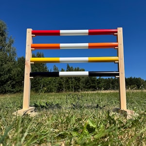 May include: A wooden horse jump with four coloured poles: red, orange, yellow, and black, each with a white stripe. The jump is set up on a grassy field with a clear blue sky in the background. The poles are approximately 152 cm long.