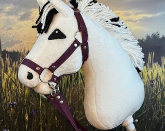 Op de afbeelding: Een wit pluche stokpaard met een paarse teugel en teugels. Het paard heeft een zwart-witte manen en staart, en een houten stok. Het speelgoed is ontworpen voor kinderen.
