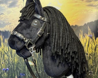May include: A black hobby horse with a plush head, featuring a black mane and a leather bridle. The wooden stick is attached to the head with a white band. The background shows a field of wheat and blue flowers.