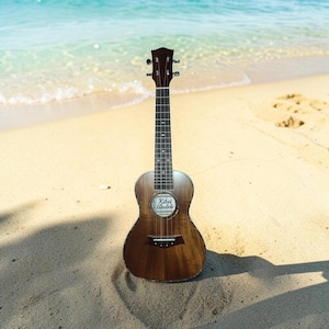 May include: A brown wooden ukulele with a dark fretboard and silver tuning pegs. The sound hole features a circular label with the text Kihei Ukulele printed in black. The instrument has four strings and a bridge positioned on the lower body.