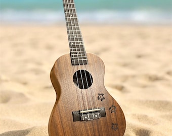 May include: A wooden ukulele with a natural finish sits on a sandy beach with the sea in the background. The instrument features a round sound hole and turtle designs. The ukulele's strings and fretboard are visible.