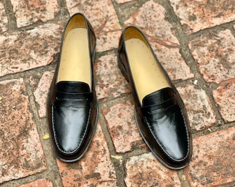 Pode incluir: Um par de mocassins de couro preto com um design clássico de tira com fenda no peito do pé, bico arredondado e salto baixo. O interior dos sapatos é forrado com um material bege claro e o exterior tem um acabamento liso e polido.
