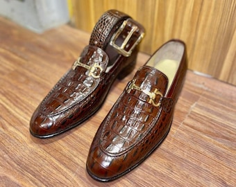 Pode incluir: Um par de mocassins castanhos com uma textura brilhante de relevo de crocodilo e um detalhe de bridão em metal dourado no peito do pé. Um sapato está na vertical enquanto o outro está inclinado, mostrando a silhueta estruturada e o acabamento polido.