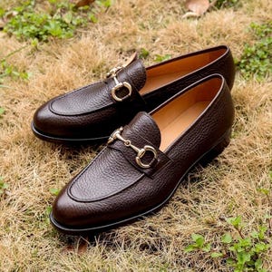 Pode incluir: Um par de mocassins de couro castanho escuro com um acabamento texturizado de gr&atilde;o e um detalhe de brid&atilde;o em metal dourado no peito do p&eacute;. Os sapatos t&ecirc;m biqueira arredondada, salto baixo e forro interior liso em bege, estilo slip-on.