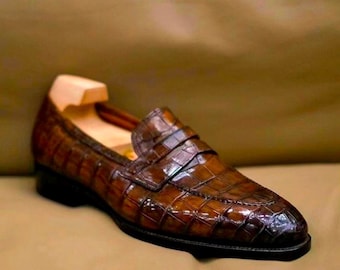 Pode incluir: Um mocassim de couro castanho com textura brilhante de pele de crocodilo, apresentando uma tira clássica sobre o peito do pé, biqueira arredondada, um salto baixo preto e uma forma de madeira clara inserida para manter a forma do calçado.