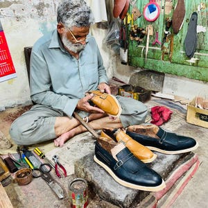 Pode incluir: Um par de mocassins de couro preto com tira de fivela e um mocassim de couro castanho claro em processo de fabrico. Os sapatos apresentam uma biqueira pontiaguda, uma sola de cor clara e um detalhe de franjas na parte superior.
