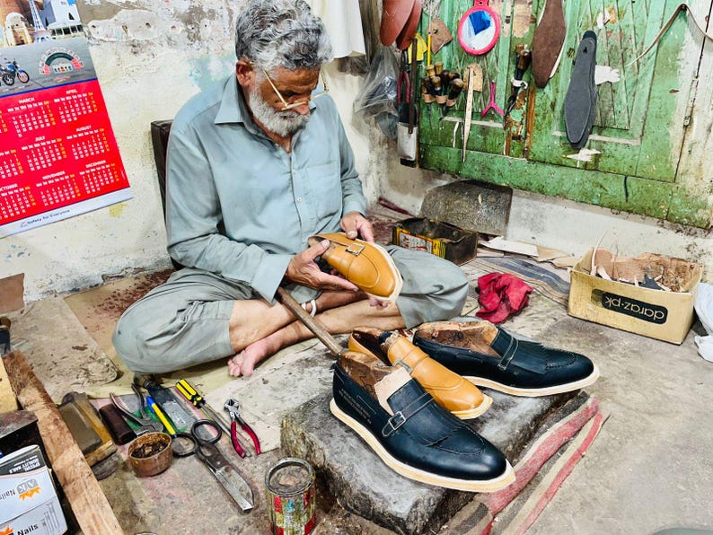 Pode incluir: Um par de mocassins de couro preto com tira de fivela e um mocassim de couro castanho claro em processo de fabrico. Os sapatos apresentam uma biqueira pontiaguda, uma sola de cor clara e um detalhe de franjas na parte superior.