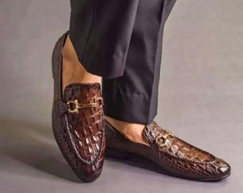 Pode incluir: Mocassins de couro castanho com textura de crocodilo. Os sapatos têm uma fivela dourada e são usados com calças pretas. Os mocassins são um estilo clássico, adequado para ocasiões formais ou semi-formais.