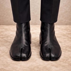 Peut inclure: Une paire de bottines en cuir noir présentant un design distinctif à bout fendu, une finition lisse et un petit talon. Les bottines sont portées avec un pantalon noir qui descend jusqu'au niveau de la cheville, révélant la couture verticale.
