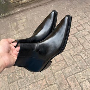 Könnte beinhalten: Ein Paar schwarze Lederstiefeletten mit polierter Oberfläche, spitzer Zehenpartie und einer markanten, quadratisch geformten Rahmennaht. Die Stiefel haben ein glattes Obermaterial, einen niedrigen Absatz und eine klare, schlichte Silhouette.