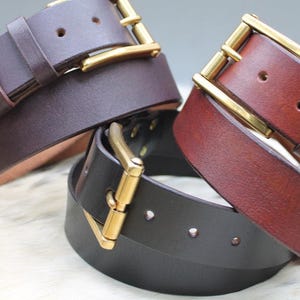 Pode incluir: Tr&ecirc;s cintos de couro nas cores castanho escuro, castanho m&eacute;dio e preto, cada um com um acabamento liso e uma fivela de metal dourado polido. Os cintos est&atilde;o dispostos em grupo, exibindo os v&aacute;rios tons de couro e as ferragens met&aacute;licas.