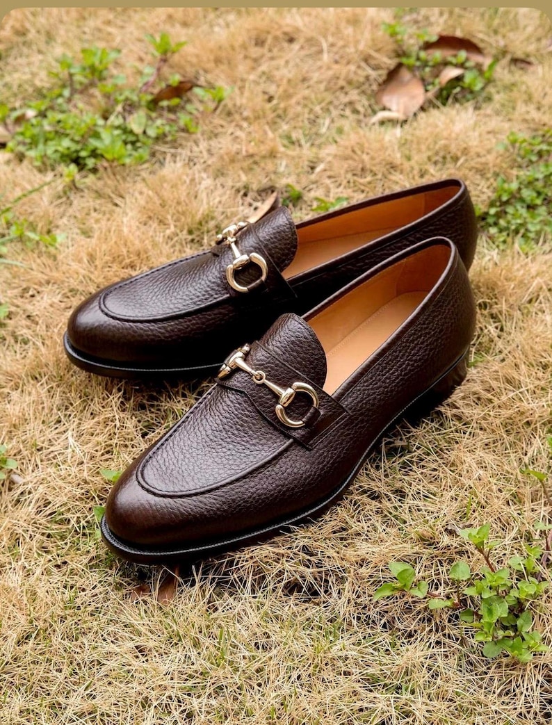 Pode incluir: Um par de mocassins de couro texturizado em castanho escuro com um detalhe de brid&atilde;o met&aacute;lico em tom dourado no peito do p&eacute;. Os sapatos t&ecirc;m biqueira arredondada, salto baixo e forro interior liso em bege, num design cl&aacute;ssico.