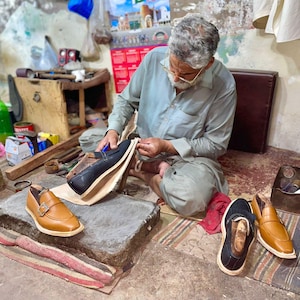 Pode incluir: Um sapateiro trabalha num mocassim de couro preto enquanto dois mocassins acabados, um castanho claro com fivela e um preto, repousam sobre um bloco de pedra. A oficina est&aacute; cheia de retalhos de couro, ferramentas e componentes de cal&ccedil;ado.