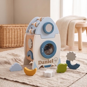 May include: A wooden rocket-shaped activity center featuring the name Daniel printed on the front. The toy includes a circular mirror, a bead maze, and various geometric shapes like a cloud, moon, star, and planet in blue, yellow, and green.