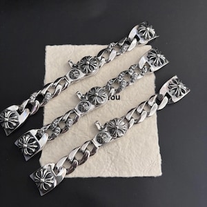 Op de afbeelding: Drie zilverkleurige kettingarmbanden met sierlijke bloemen- en kruisontwerpen. De armbanden hebben een dikke gourmettekettingstijl en worden gepresenteerd op een neutraal gekleurd oppervlak tegen een donkere achtergrond. Elke armband heeft een decoratieve sluiting.
