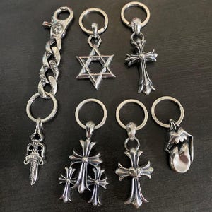 May include: A selection of silver keyrings with diverse designs, including crosses, a six-pointed star, a dagger, and a tongue. One keyring features a chain link design. The keyrings are displayed on a dark surface.