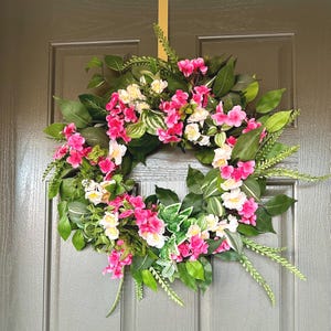 May include: A pink and white floral wreath with green leaves. The wreath is hanging on a gray door.