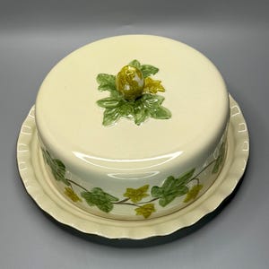 May include: A cream-colored ceramic butter dish with a lid. The lid is topped with a green and yellow floral design. The base of the dish is decorated with green ivy leaves. The dish has a scalloped edge.