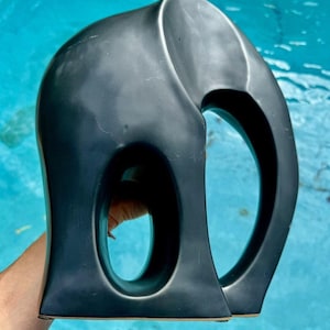 May include: A dark gray ceramic elephant sculpture with a smooth, matte finish. The sculpture has a stylized design with two large, oval openings. The elephant is held against a blue water background.