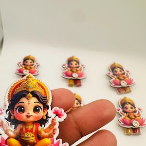 Puede incluir: Colección de figuras decorativas de madera coloridas. Cada figura representa una deidad de estilo caricaturesco sentada sobre una flor de loto rosa, adornada con joyas doradas y una pieza central enjoyada. Las figuras están sobre un fondo blanco.