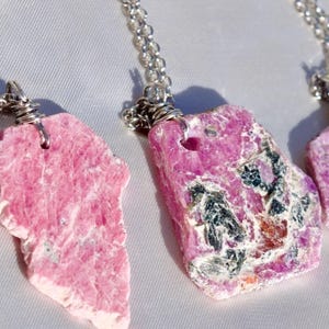 Puede incluir: Tres colgantes de piedras preciosas rosas y moradas en cadenas de plata. Los colgantes están tallados en bruto y tienen un aspecto natural y orgánico.