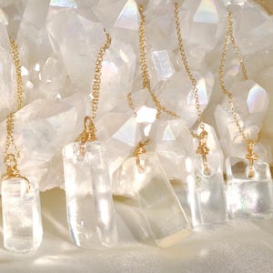 May include: Clear quartz crystal necklaces with gold-colored chains and accents. The rectangular pendants hang in front of a backdrop of large, white quartz crystals. The necklaces are displayed on a white, silky surface.