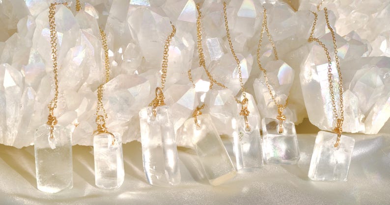 May include: Clear quartz crystal necklaces with gold-colored chains and accents. The rectangular pendants hang in front of a backdrop of large, white quartz crystals. The necklaces are displayed on a white, silky surface.