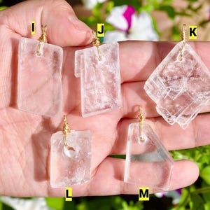 May include: Five clear, rectangular and geometric pendants with gold-colored wire loops. The pendants are held in a hand, with a blurred background of green foliage and white and purple flowers.