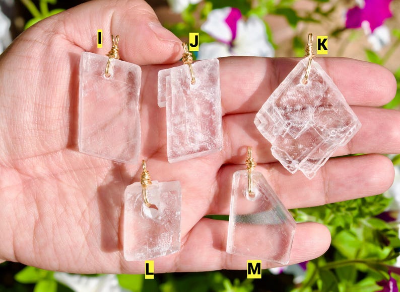 May include: Five clear, rectangular and geometric pendants with gold-colored wire loops. The pendants are held in a hand, with a blurred background of green foliage and white and purple flowers.