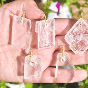 May include: Five clear, rectangular and diamond-shaped pendants with gold-colored wire loops. The pendants are held in a hand, with a blurred background of green foliage and flowers.