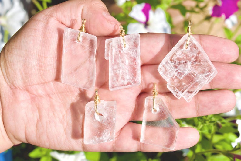 May include: Five clear, rectangular and diamond-shaped pendants with gold-colored wire loops. The pendants are held in a hand, with a blurred background of green foliage and flowers.