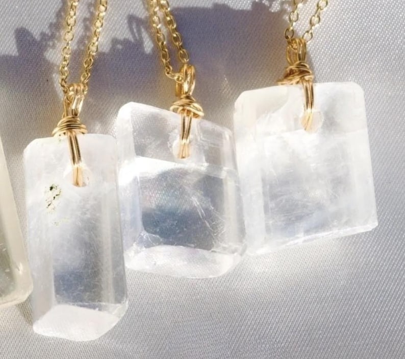 May include: Close-up of three rectangular, translucent white selenite crystal pendants. Each crystal is wire-wrapped with gold-colored metal and suspended from a delicate gold chain. The pendants are displayed against a soft, light-colored background.