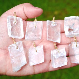 May include: Eight clear, rectangular pendants with gold-colored wire loops are displayed in a hand. The pendants are made of a translucent, crystalline material. The background is a blurred green.