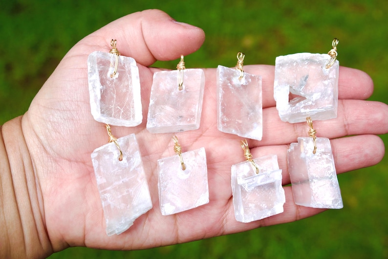 May include: Eight clear, rectangular pendants with gold-colored wire loops are displayed in a hand. The pendants are made of a translucent, crystalline material. The background is a blurred green.