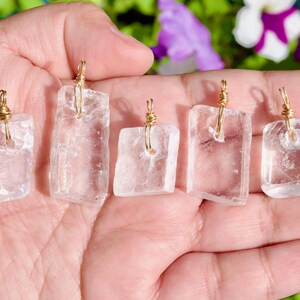 May include: Five clear quartz crystal pendants with gold-colored wire wrapping. Each pendant is a different rectangular shape. The crystals are held in an open hand against a blurred background of green and purple.