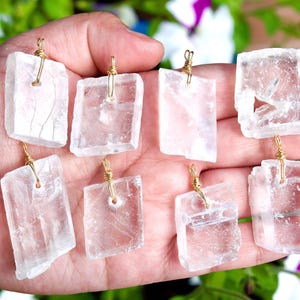 May include: Eight clear, rectangular crystal pendants with gold-colored wire loops are held in a hand. The crystals have a rough, natural texture and varying shapes. The background is blurred with green and purple foliage.