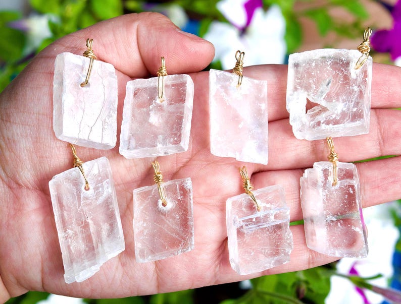 May include: Eight clear, rectangular crystal pendants with gold-colored wire loops are held in a hand. The crystals have a rough, natural texture and varying shapes. The background is blurred with green and purple foliage.