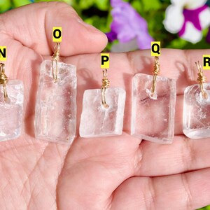 May include: Five clear quartz pendants with gold wire wrapping. Each pendant is rectangular with slightly irregular edges. The pendants are labeled N, O, P, Q, and R with yellow stickers. The background includes green foliage and purple flowers.