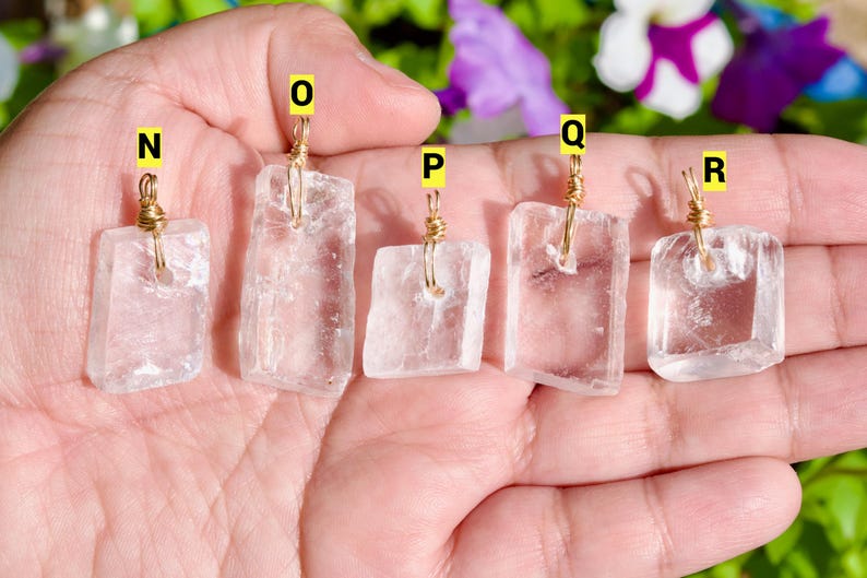 May include: Five clear quartz pendants with gold wire wrapping. Each pendant is rectangular with slightly irregular edges. The pendants are labeled N, O, P, Q, and R with yellow stickers. The background includes green foliage and purple flowers.